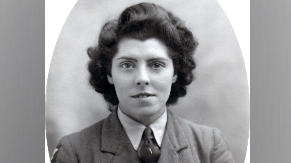 A young woman with dark, short curly hair, smiles at the camera, wearing an army uniform.