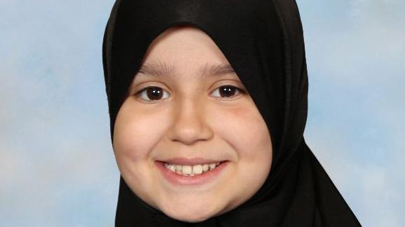 A photo of Sara Sharif smiling wearing a black head scarf. She is looking at the camera and in front of a blue background