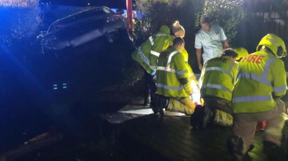 A silver VW Passat poking through a hedge it has crashed through.  In the foreground are firefighters