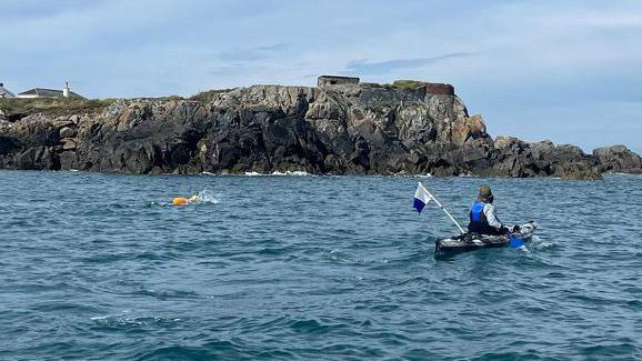 Rosie Williams can be seen swimming in the sea off the rocky coastline while a kayaker is a short distance away from her.