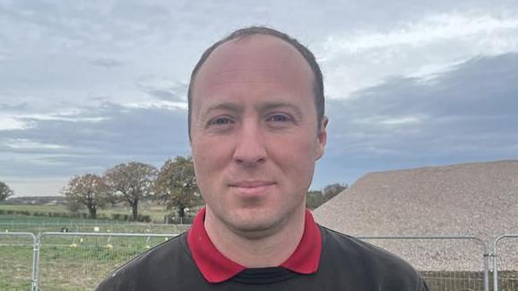 David Boden smiles while standing outside in a field on a grey day. He has short dark hair and wears a dark green jumper with a red polo top underneath.