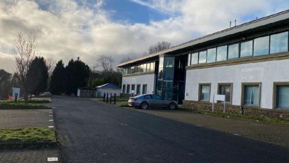 A white building with a row of windows on the top floor. There is a road in front of it and a row of spaces for parking with one grey car parked up.