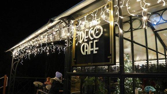 The exterior of The Heritage Deco Café on a winter's night. There are strings of fairy lights hanging from the exterior of the temporary structure, in front of the art-deco sign. A woman and a child can be seen wrapped up warm as they sit outside.