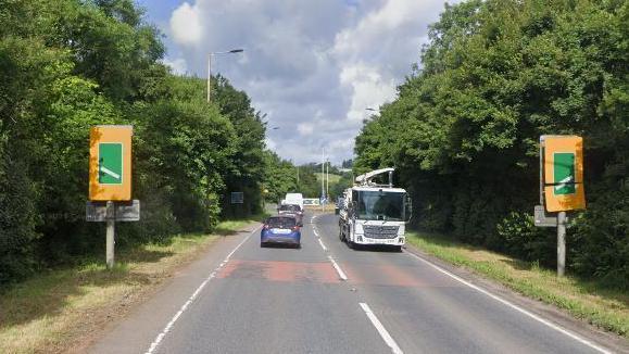 Cars, vans and a lorry drive along the A361 in Bish Mill near South Molton in Devon. The vehicles are approaching a roundabout with signs on both sides of the road showing how close to the roundabout they are. Trees line both carriageways and it is a cloudy day.