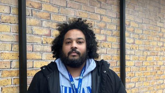 A man stands in front of a brick wall at Chelmsford station. He has black curly hair and a black beard. He is wearing a grey hoody and black jacket. 