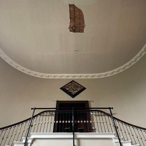 Plaster has fallen away from a white church roof, revealing wooden lathes on the ceiling. Below is a pair of dark wood double doors, and two sets of black staircases - one on the right and one on the left