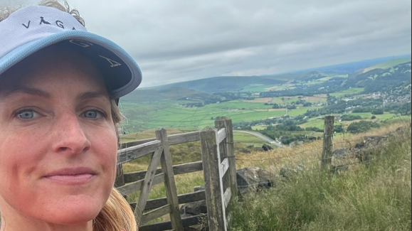 A woman with blonde hair standing in front of a gate overlooking hills an fields 