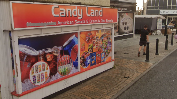 The shopfront of Candy Land. It has an orange banner that advertises its name, as well as pictures of various goods including fizzy drinks, crisps and sweets. The shop is in Braintree town centre. Other shops are pictured in the background.