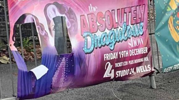 A photo of the defaced poster, you can read the writing but there are two holes cut out where the artists faces should be. It is clearly a poster attached to railings on a pavement.