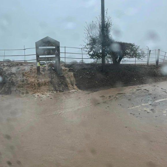 A view from inside a car of water and mud running down a small verge, underneath a road sign. There is mud and water covering the road.