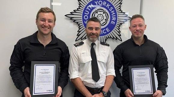 PC Ben Hawley (left) and PC Carl Holden (right) pictured, smiling and holding their awards, with Doncaster District Commander Ch Supt Pete Thorp (Middle) in front of the South Yorkshire Police emblem.