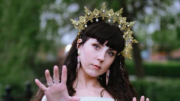 Shannon Pearl. She has long dark hair and is wearing a white dress. She is wearing a gold tiara which is decorated with pearls. She has an intricate white swirl painted between her eyes and a white line running from her bottom lip to her chin.