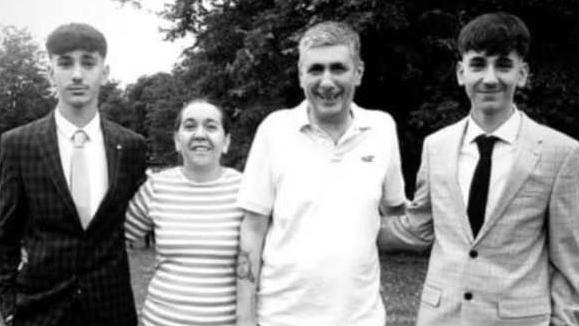 The family of four are standing outside with their arms around each other. They are all facing the camera. There are three men, two siblings and and older man, who are a similar height. Between them is a women who is much smaller. They are all smiling and the picture has been taken in black and white.