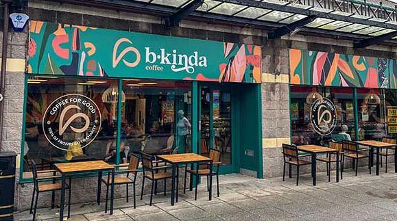 The outside of b.kinda coffee shop on the Ridgeway. The shop is glass fronted with green geometric signage. There are wooden tables and chairs outside the front. 