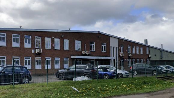 An outside view of the two storey Permoid Industries factory in Newton Aycliffe with a row of cars parked outside. The factory is made of brick with white windows. The roof rises to a central point at a shallow angle.