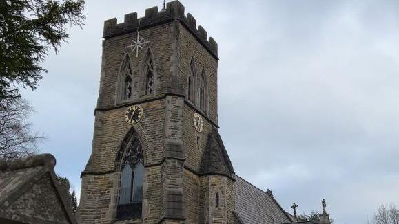 Image shows a church with a stone tower and arched windows with a black and gold clock with reads 12:35. The sky is grey and cloudy. 