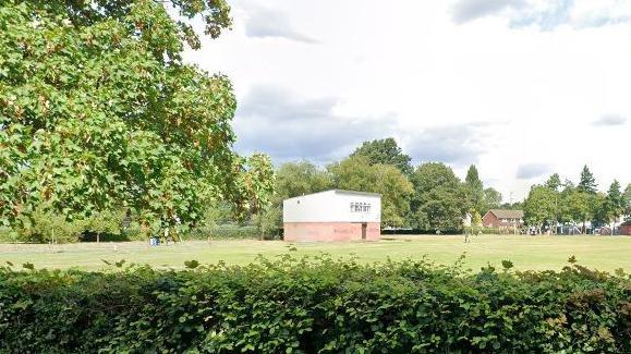 A Google Streetview image of the pavilion which is a low building with a red brick lower part and white rendered upper part. The pavilion is in the middle of a park with grass around it and trees behind it, while there is a low hedge in the foreground of the picture.