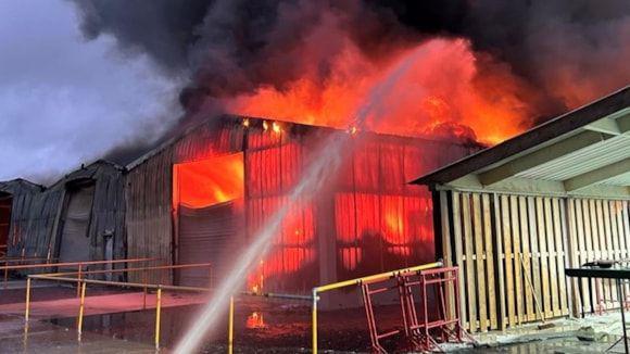 A building up in orange and red blazes, with big black smoke plume above. To the bottom, we can see water being thrown in the direction of the building. 