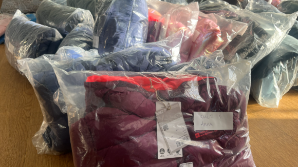 The photo shows a pile of new clothing in clear bags with labels on the floor.