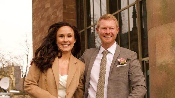 A woman with long dark hair looks at the camera next to a man with short, red-blonde hair. She wears a beige jacket over a white top while he wears a grey suit jacket with a pink flower in his pocket and a grey patterned tie against a white shirt. Behind them are the stone walls and columns of the cathedral.