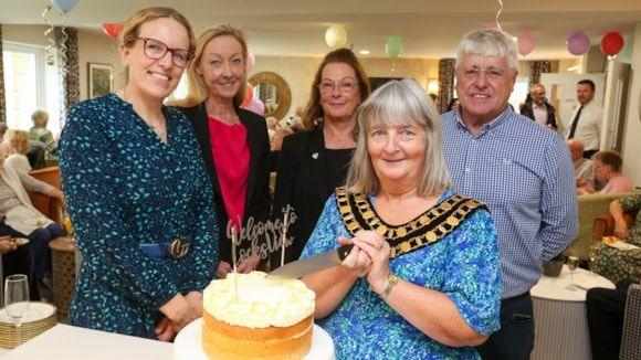 Five people (four women and a man) surrounding a cake and smiling at the camera. One woman has mayoral chains on. There are balloons in the background and people milling about.