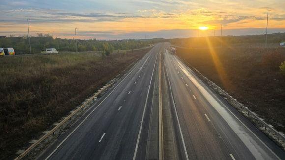 A picture of the A421 carriageway. The photo is taken from a bridge over the carriageway. The road is clear and traffic can be seen heading onto the road via a slip road. The sun can be seen rising in the distance. 