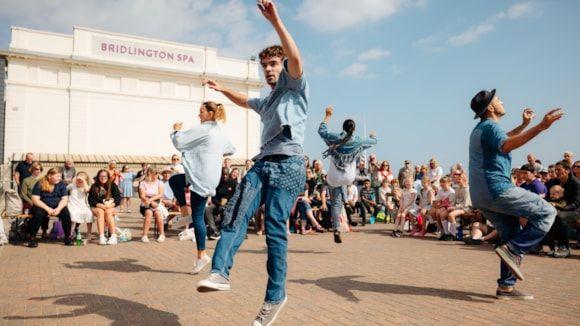 Four street performers, made up of two men and two women wearing denim jeans and denim trousers, are performing a dance on the street outside a theatre with people sat down watching them.