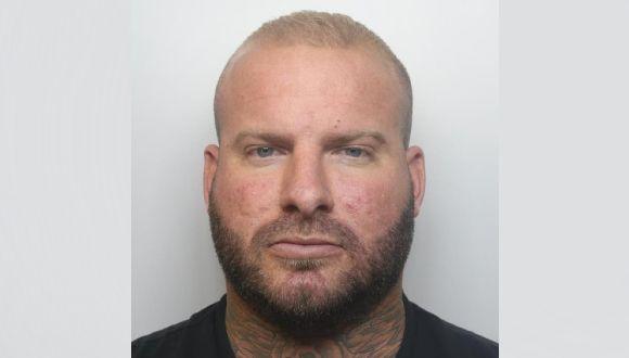 A mugshot of Alex Male standing against a grey wall. He is wearing a black t-shirt and short blonde hair.