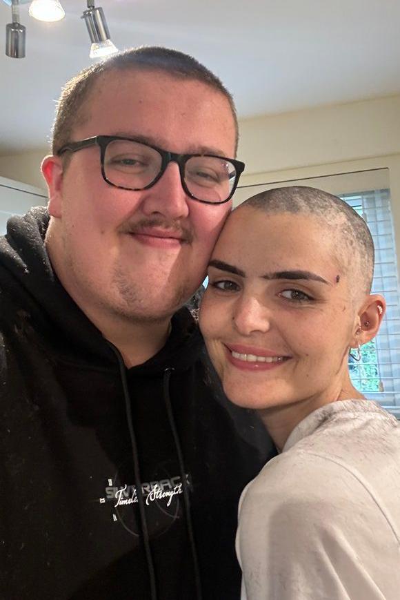 A man and a woman smiling together. The woman has a shaved head.