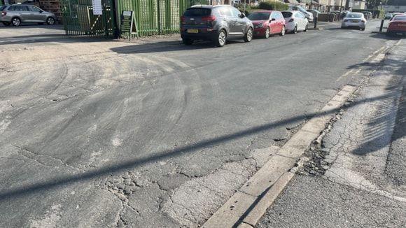 View of Seavy Road showing cracks in the tarmac on the path and road. There are  cars parked along one side of the road, as well as an entrance gate to premises.