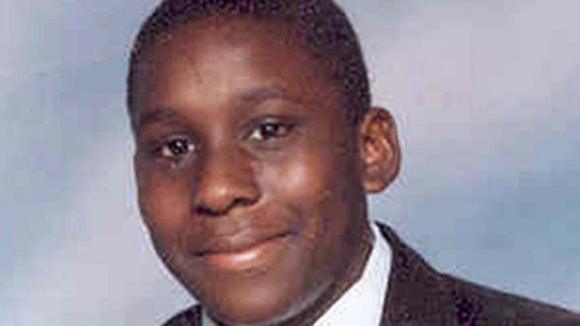 Anthony Walker is pictured wearing his school uniform, including a black blazer and white shirt, in this professional school photograph. There is a cloudy blue and white background and he is smiling at the camera.
