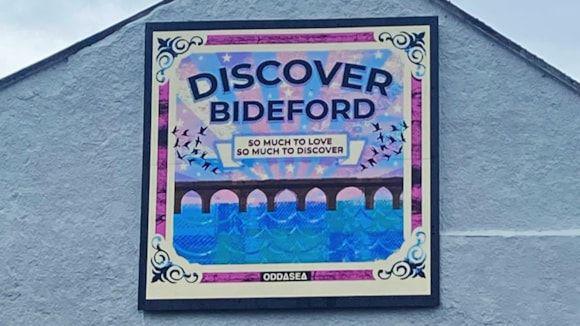 The 2m by 2m artwork sits at the gable end of Country Cottage Bakery. It is a white background with a bridge, water and DISCOVER BIDEFORD - written above it in all capital letters. There are purple and blue stripes in the design.