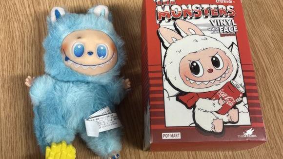 A doll and its box, with a wooden background. The blue doll is on the left, with blue eyes and a spiky grin. The red packaging is on the right, advertising a "vinyl face blind box".