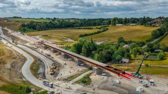 Section of railway route cutting a diagonal swathe across the countryside. The narrow dark red deck is seen along the route, supported by concrete pillars. There are various vehicles at the construction site, and two temporary access roads are on the right of the deck. There are trees and fields in the surrounding area.