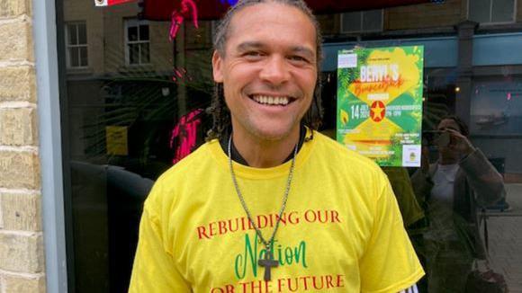 A smiling Lee Quashie wears a yellow shirt, reading "rebuilding our nation for the future", and a necklace with cross.