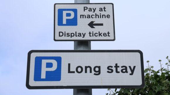 Two signs on a lamppost. The first is a white square sign with a black border. It has a blue square with a white P in. Next to it on the right, it reads "Pay at machine" with a black arrow pointing left. Below, it reads "Display ticket". The second sign which sits below the first is a rectangular white sign with a black boarder. It also has a blue square with a white P. Next to it on the right it reads "Long Stay". 