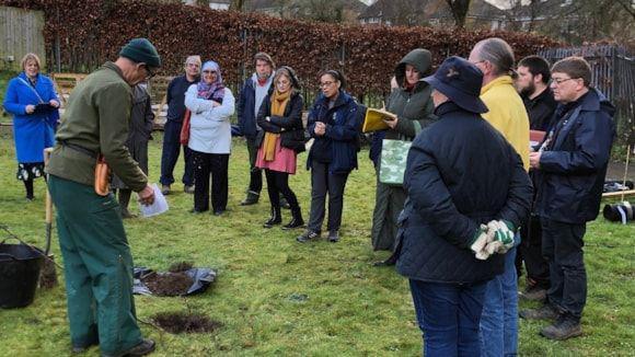 A wide shot photo of 12 people stood on a cold winter day. They are watching a man demonstrate how to plant a tree.