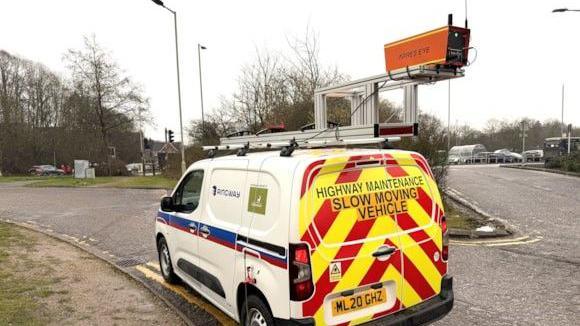 An AI "camera" is sitting on the top of a council road maintenance van. The scanner is orange and is fixed on using a steel structure. The van is white and has red and yellow chevrons at the back and the council logo on the side. It is driving along a road