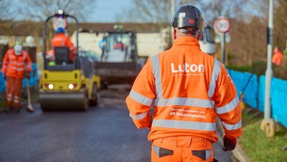 A man wearing bright orange high-vis clothing is walking towards some roadworks ahead of him. On his back the words "Luton" are written in white font.