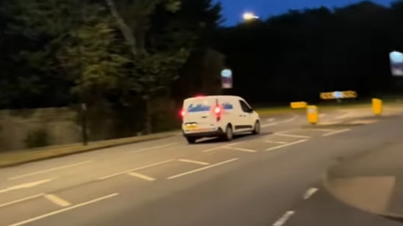 A screenshot from a video of a white van with blue writing and red brake lights on driving towards a roundabout in Coventry