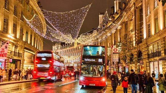 Red London buses travelling up and down Regent Street at night, with Christmas lights overhead. The 88 bus is at a bus stop on the right and there are pedestrians on the pavements on both sides of the street. 