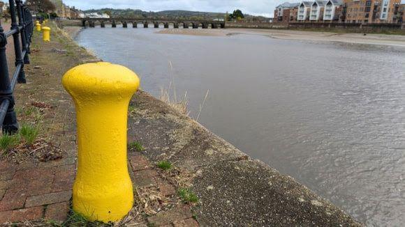 Castle Quay at Barnstaple on a cloudy day. There is a row of yellow mooring bollards along the iron railing of the quayside with one prominent in the foreground. Over the calm water is a row of five-storey housing. Spanning the water in the background is a road bridge.