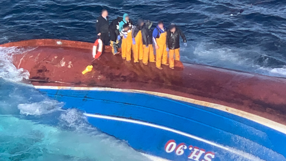 The crew of the Njord standing on the upturned hull of the boat. They are all wearing yellow fishing waders and are standing in a line on the red part of the hull. Several are wearing dark clothing. The bottom of the hill is red and is separated from the blue top of the hull by a white line. the letters SH.90 are upside down on the blue section.