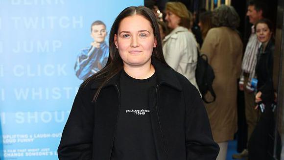 Andrew, wearing a black sweatshirt and jacket, stands on the red carpet in front of a huge film poster for I Swear at its premiere. She wears her long dark hair down.
