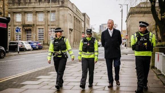 Clive Grunshaw walking with three uniformed police officers on a street lined with old stone buildings