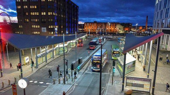 Night time shot of buses in Liverpool. People are crossing the road as buses pull into stops at the bus shelters.  Albert Dock is in the background.  There is a clock in the foreground. 