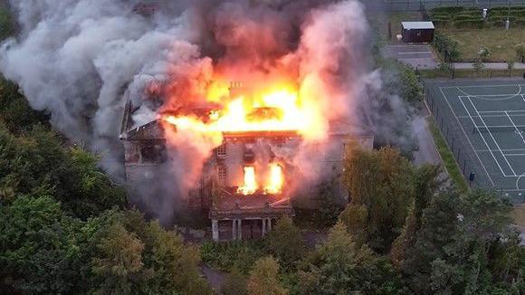 Drone footage of a two-storey 18th Century manor house engulfed in orange flames with thick grey smoke billowing into the sky