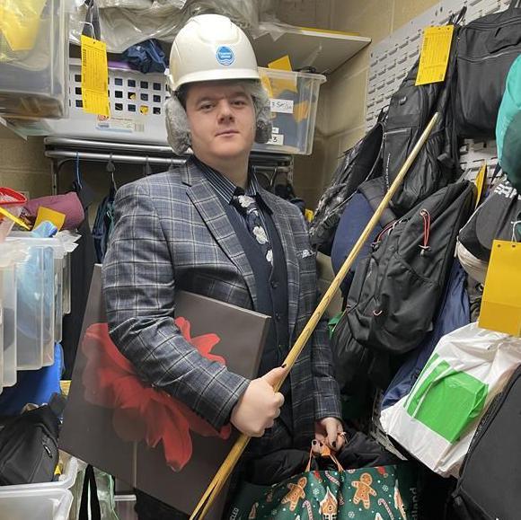 A customer service officer in a lost property storeroom with some of the lost items, including a hard hat (on his head) and snooker cue.