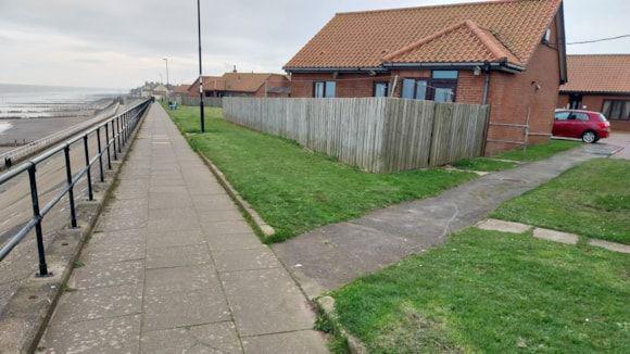 A paved coastal path beside a row of red brick houses with orange-tiled roofs. A wooden fence surrounds part of a grassy yard. Another path cuts through the lawn on the right. A black metal railing separates the walkway from the beach on the left. The sky is overcast and the sea, which can be seen on the top left hand corner of the photo is grey. 