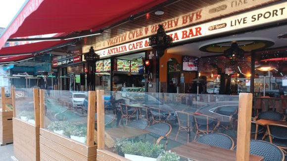 A seating area with glass panelling and wooden benches outside the front of the Antalya cafe, seen adorned with a white and red cafe sign.